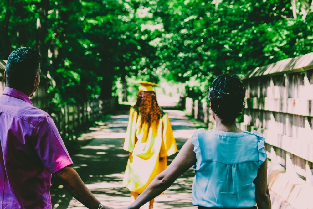 girl in yellow graduation gown and cap walking in front of parents holding hands